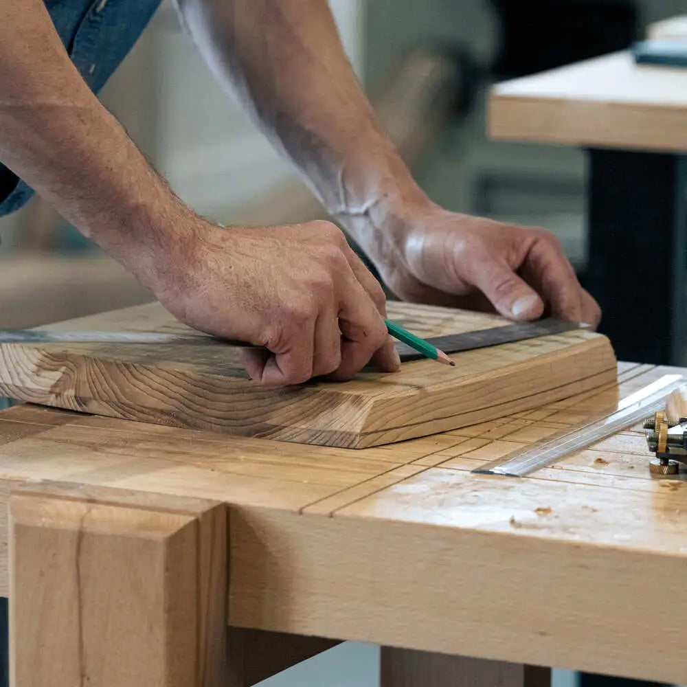 A person’s hands carefully marking a piece of light-colored wood with a green pencil and metal ruler.